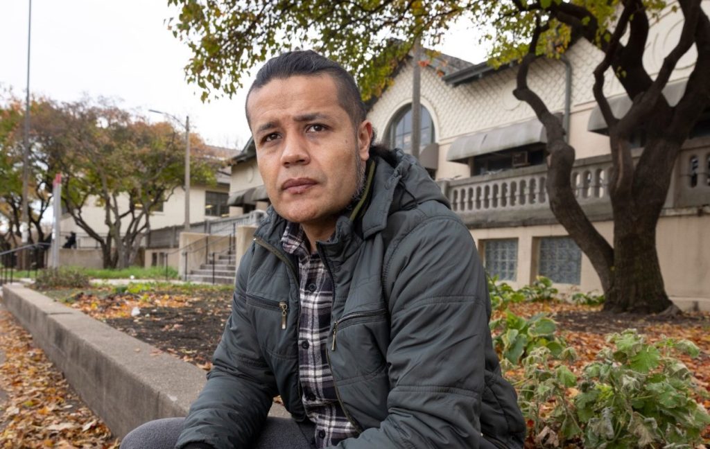 A migrant from Colombia sits in front of a building. He is wearing a gray jacket and plaid shirt. He looks off into the distance.