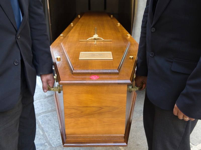Two pallbearers in dark suits carry a coffin in a close-up photo