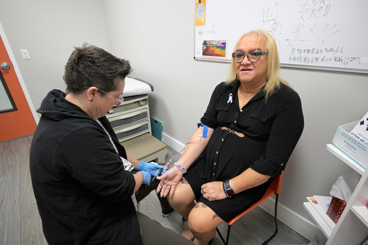A medical personnel, dressed in a black hoodie, draws blood from Andrea Montanez, dressed in a black dress, as she sits a medical office.
