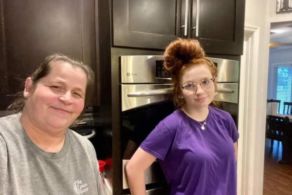 Teresa Edenfield and daughter Layken Edenfield stand in a kitchen. Teresa wears a gray t-shirt and smiles. Her hair is pulled back. Layken wears a purple ti-shirt and a necklace. Her red hair is pulled up on top of her head and she wears horned-rimmed glasses.