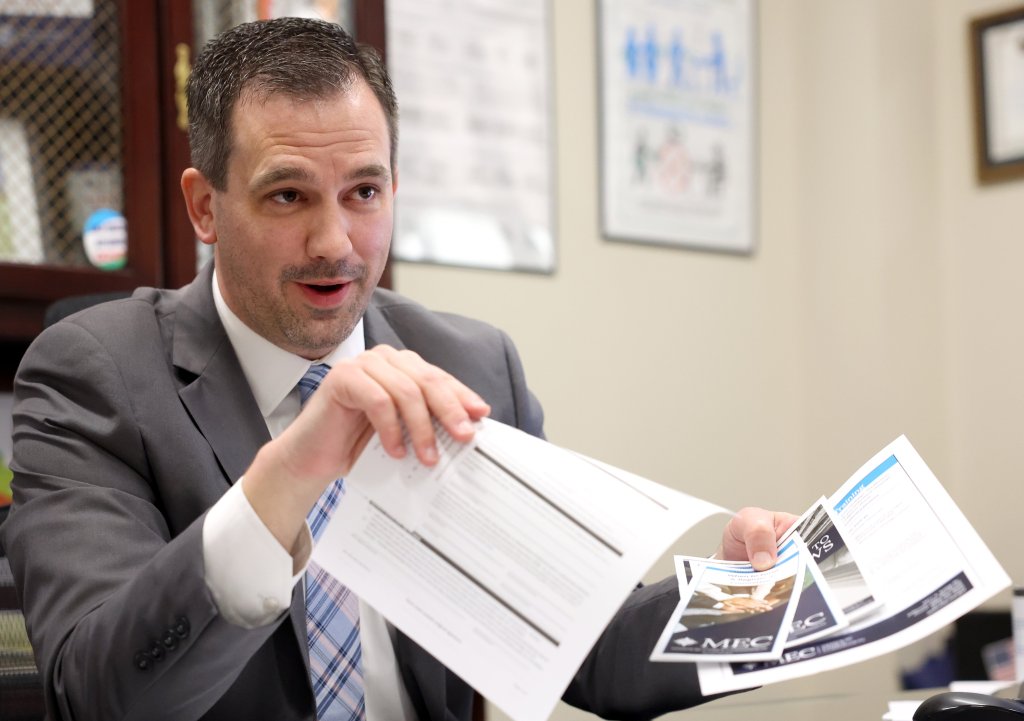 Eric Fey is holding paperwork in both hands and looking off-camera at the person he's talking to. Behind him, framed posters and other items hang on the walls.