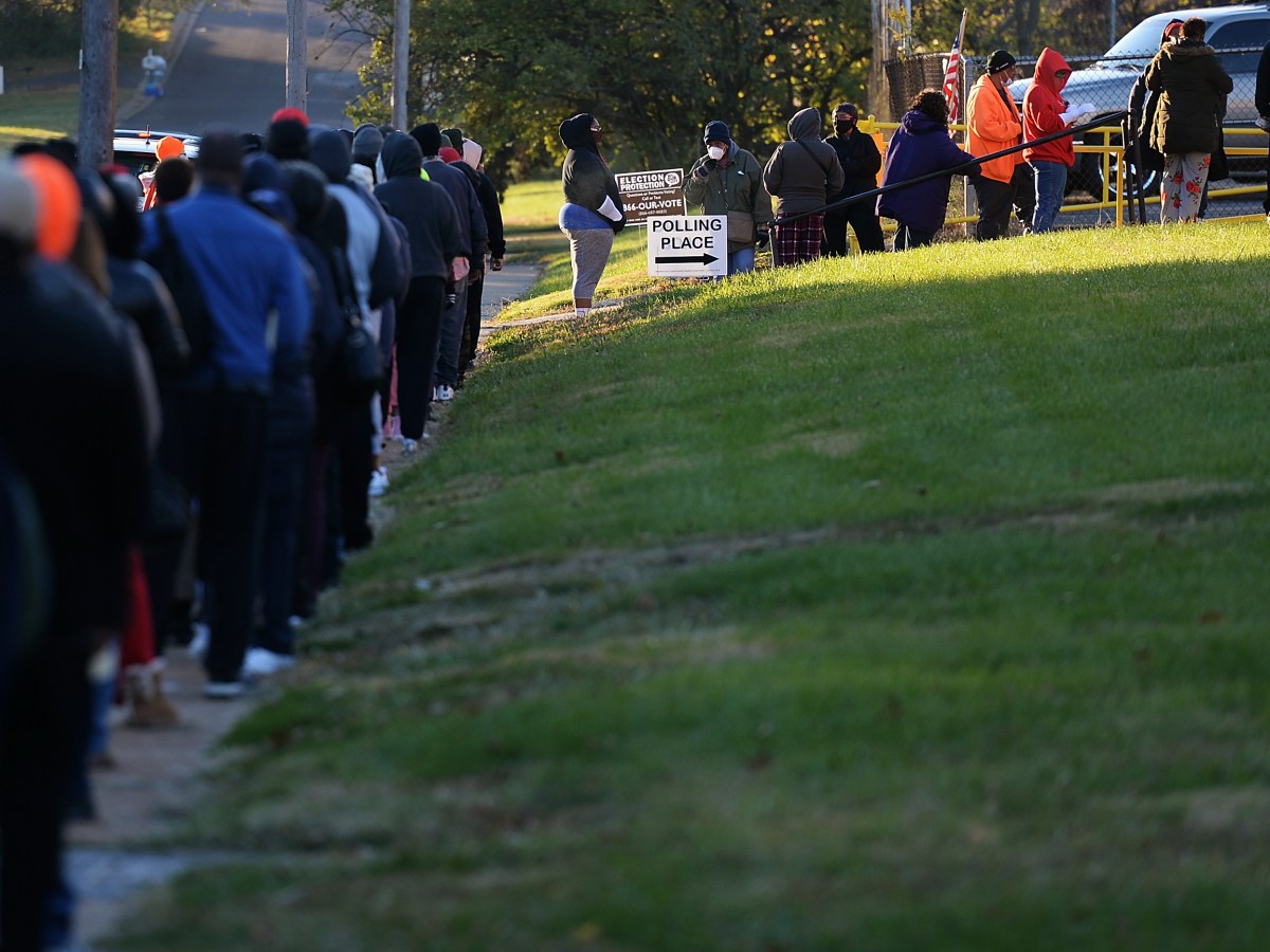 The line of voters is long, wrapping down a pathway up to the building and alongside the road. People are dressed in coats and wearing face masks. A sign says "Polling Place" with an arrow.
