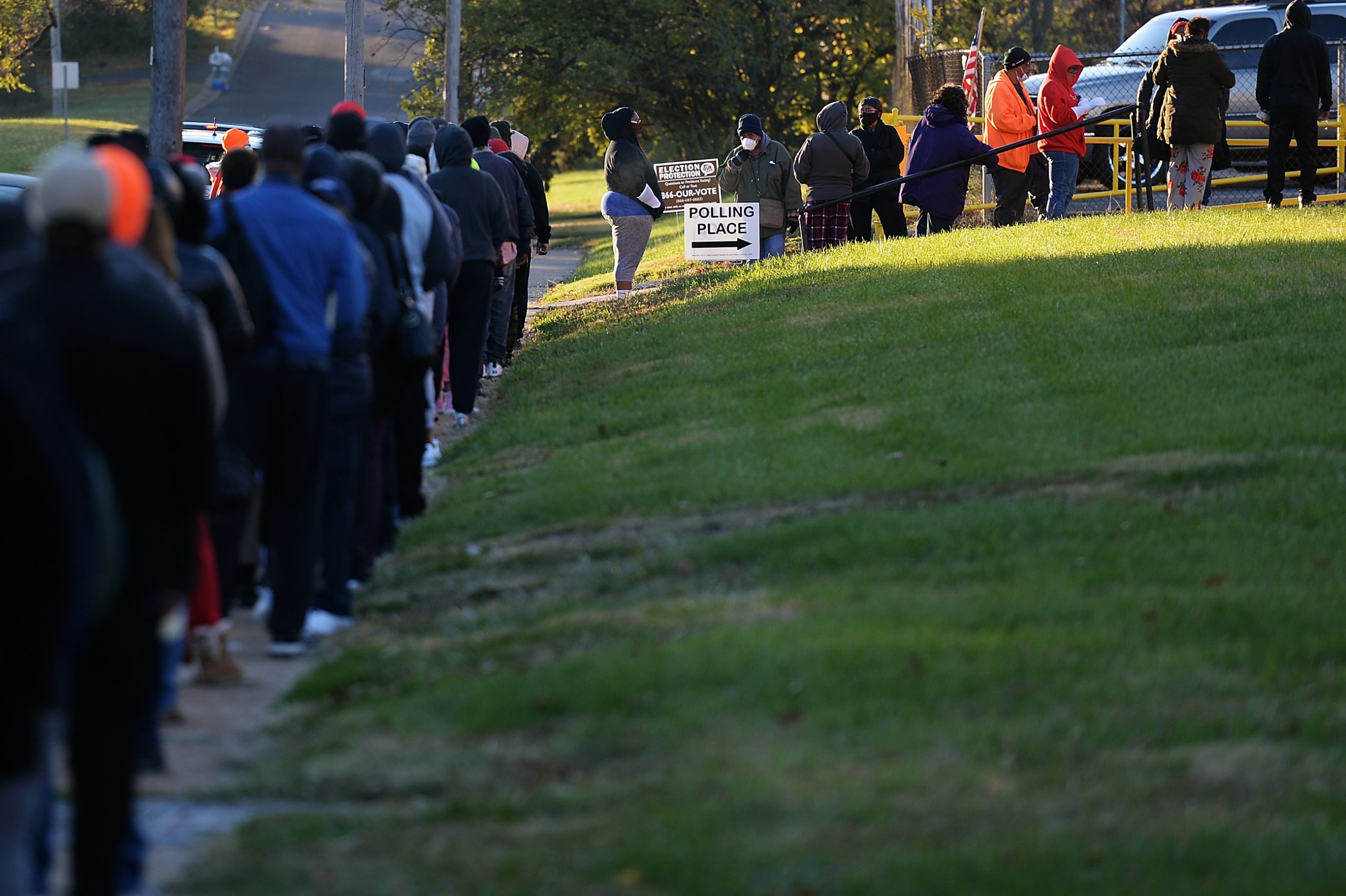 The line of voters is long, wrapping down a pathway up to the building and alongside the road. People are dressed in coats and wearing face masks. A sign says "Polling Place" with an arrow.