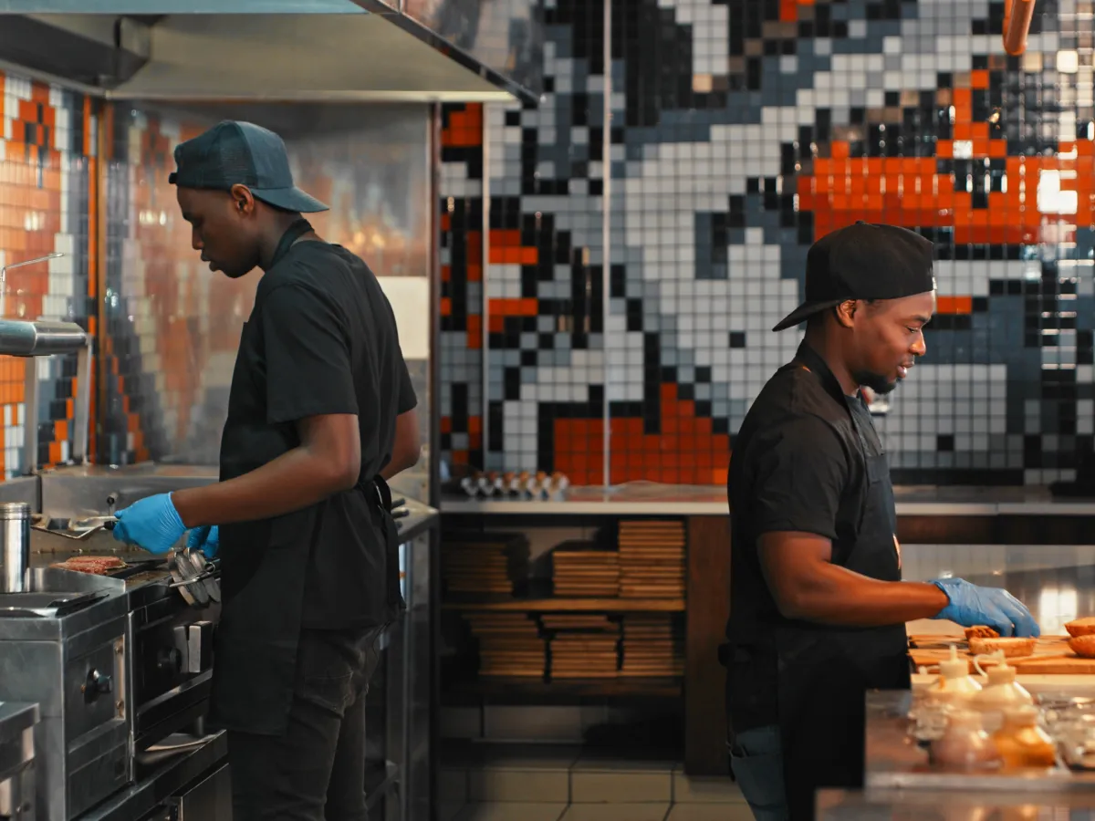 Two men work at a restaurant cooking burgers on a grill and at a prep station. Generative AI has the potential to eliminate their jobs.