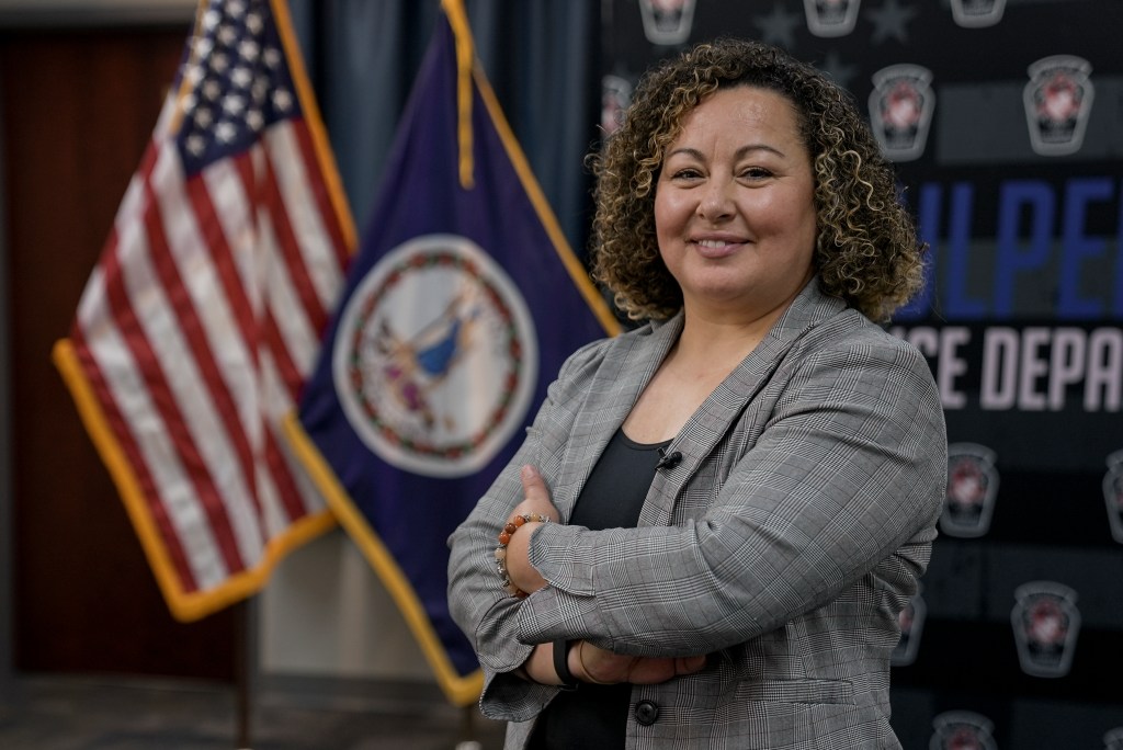 Norma McGukin wears a gray blazer with her hands folded across her chest as she poses in front of an American and Virginia flag.