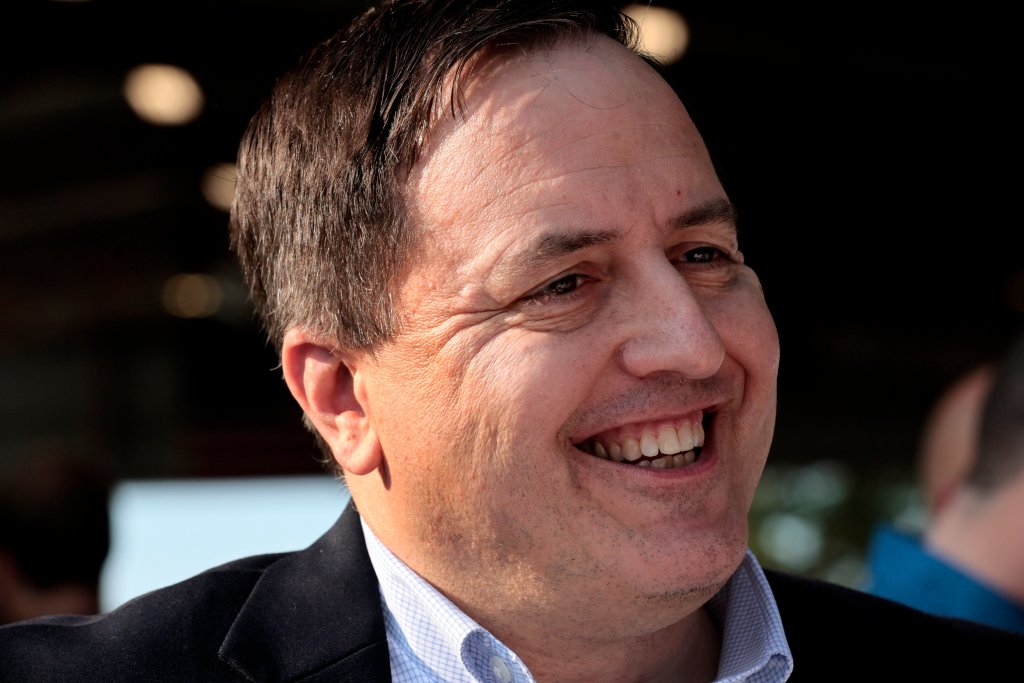 Photo shows a close-up of Jay Ashcroft, looking off-camera, smiling and wearing a checked shirt and a dark blazer.