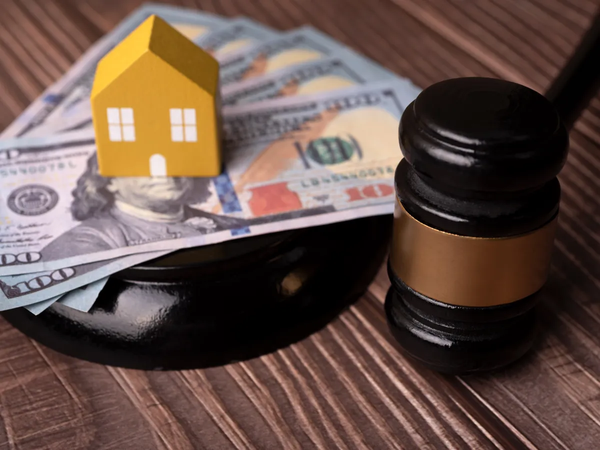 Image shows a miniature house sitting on several bills of money, next to a gavel