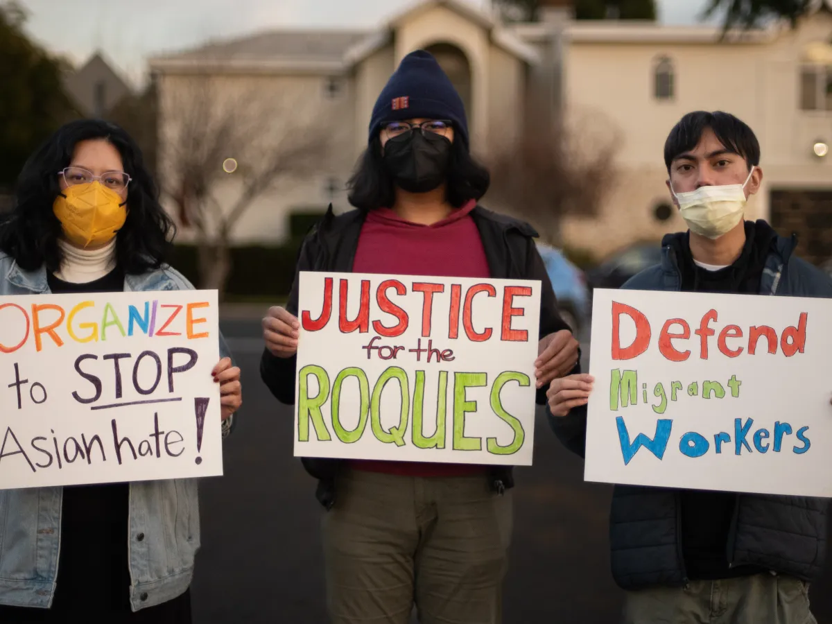Three people stand outdoors, wearing face masks to protect against COVID, and holding signs. The signs say: "Organize to STOP Asian hate!" and "Justice for the Roques" and "Defend Migrant Workers"