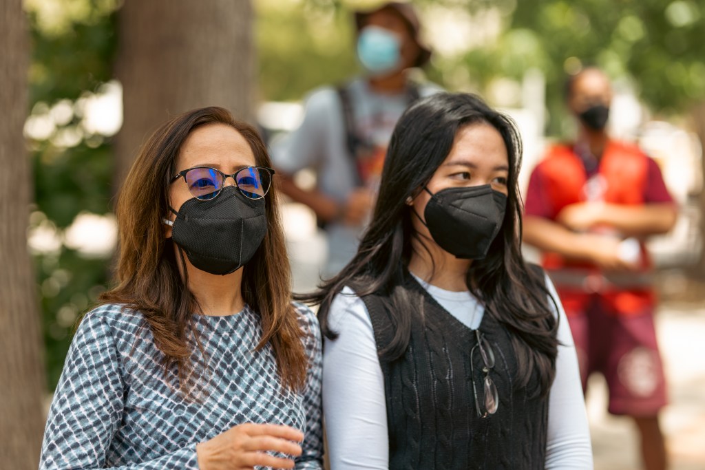 The woman are standing shoulder-to-shoulder near trees and a few other people. Everyone is wearing masks to protect against COVID.