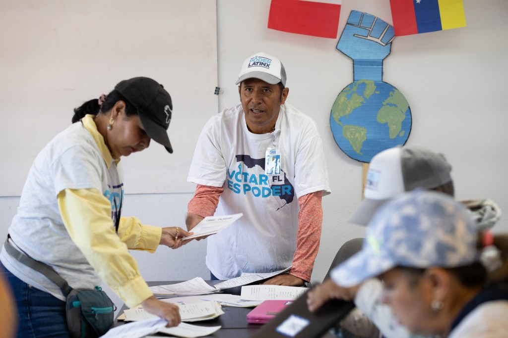 Humberto Orjuela is holding registration forms and talking to someone off-camera. People standing near him are looking at forms on a table. Behind them, there are flags on the wall and the Poder Latinx symbol.