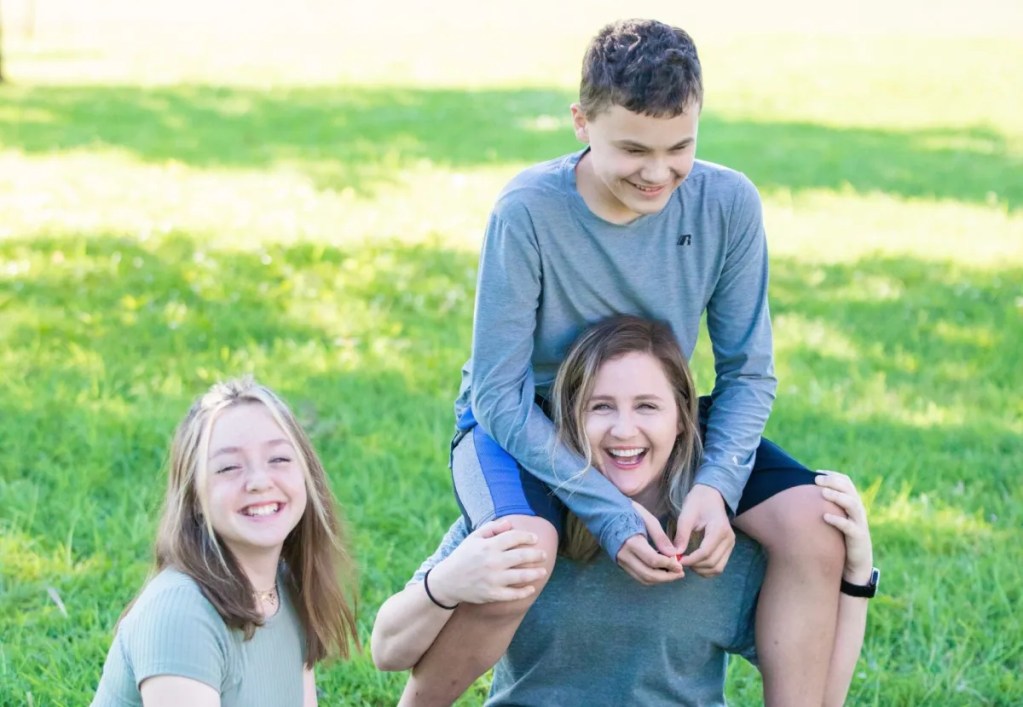 A young gift with long hair wearing a short-sleeved green shirt smiles. She stands next to a woman who is smiling with a boy on her shoulders.