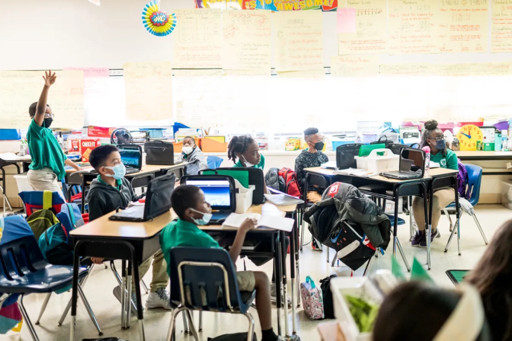 Students are in a classroom at desks. They wear green polo shirts, tan shorts and most are wearing masks. One student in the back stands with his hand raised.