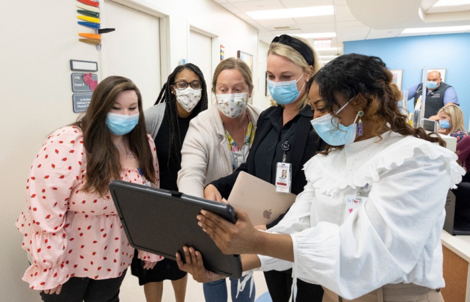 Five nursing wearing masks stand looking at a tablet. They are in a hospital with the nurses station behind them.