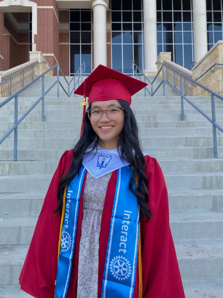 Victoria Zhang, in her graduation robe and cap, smiles on the exterior steps of the school