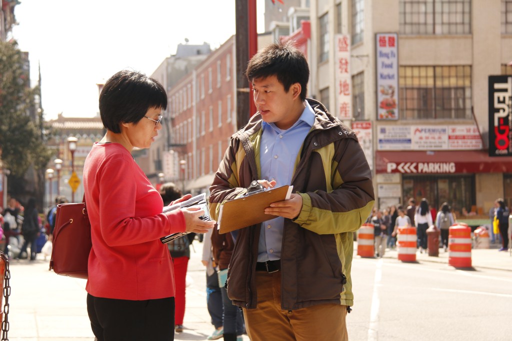 Wei Chen is holding a clipboard and talking to a woman on a sidewalk.