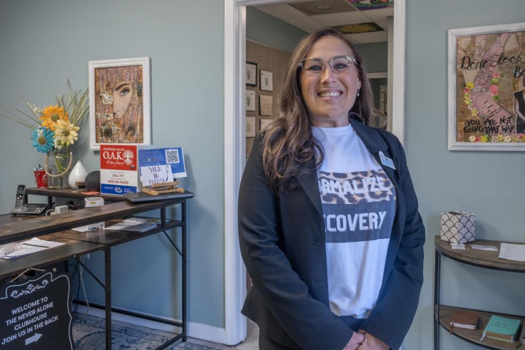 The director of the Never Alone Clubhouse stands in the lobby of the organization's office. She has long hair and glasses and is smiling. She wears a gray jacket and a t-shirt that reads Normalize Recovery. The front desk is behind her.
